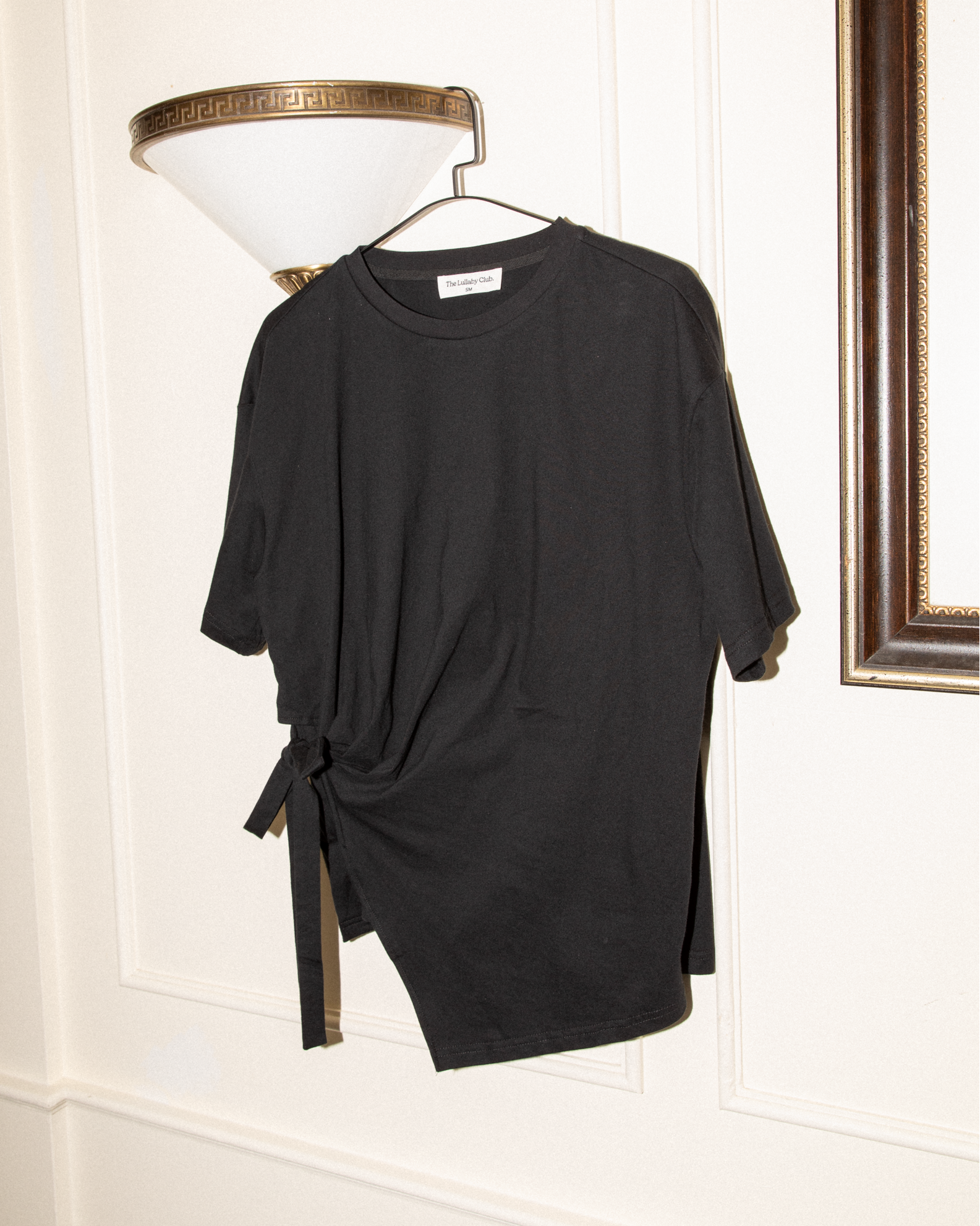 A black short-sleeve t-shirt with a side tie detail, hanging on a hanger against a white paneled wall next to a wall lamp.