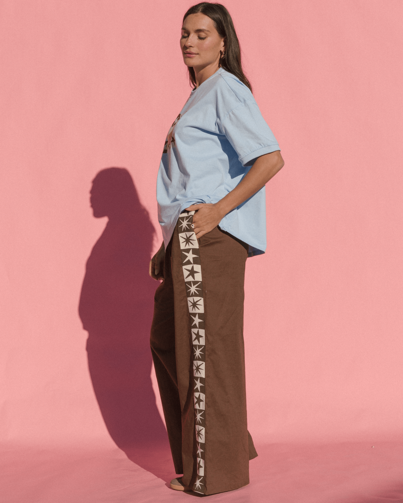 Woman wearing a light blue shirt and brown pants with a pattern against a pink background