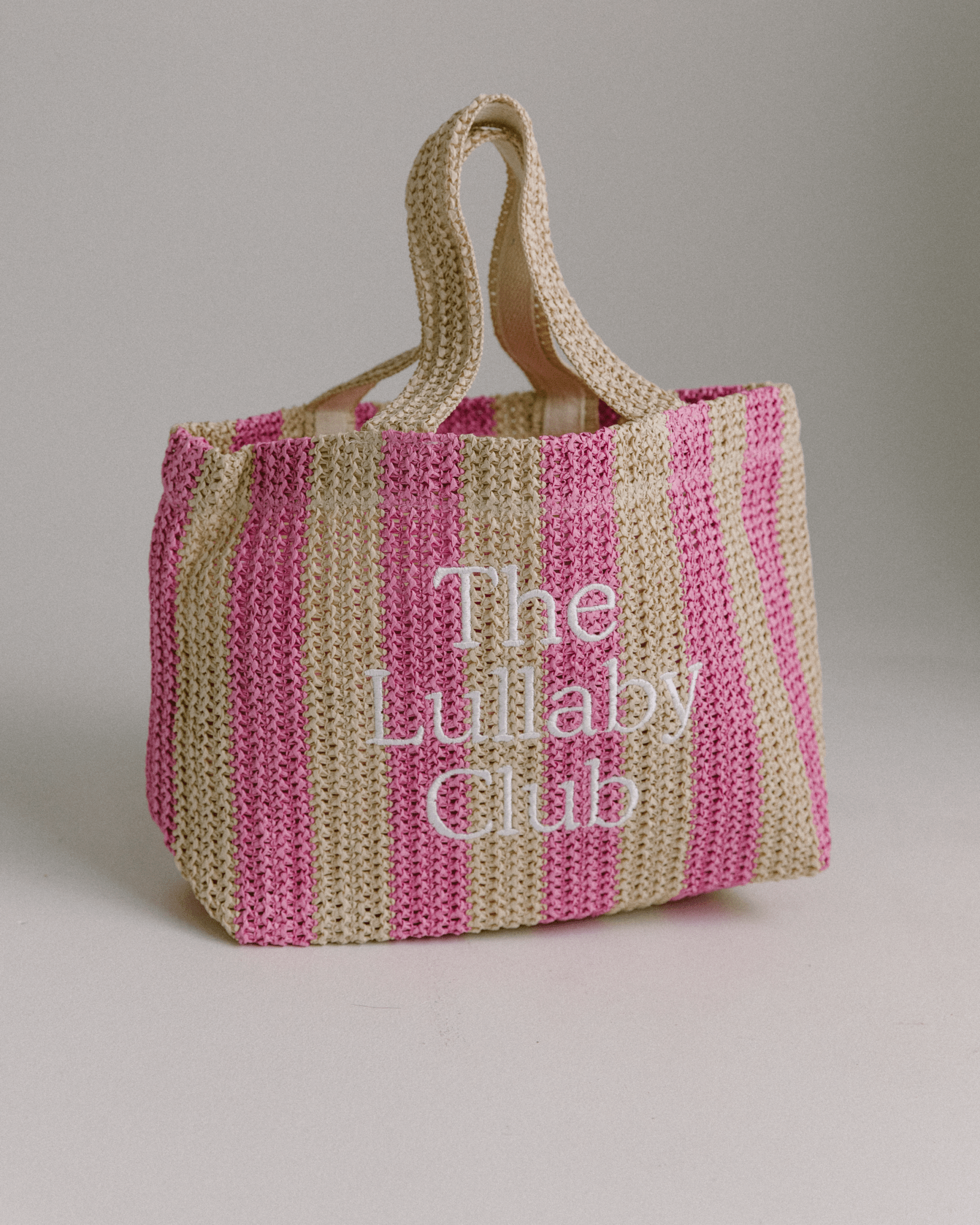 A striped pink and beige raffia tote bag with 'The Lullaby Club' embroidered on it.