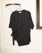A black short-sleeve t-shirt with a side tie detail, hanging on a hanger against a white paneled wall next to a wall lamp.