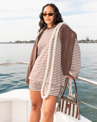 A woman stands on a boat wearing a brown and white striped long-sleeve top and matching shorts, with a brown jacket draped over her shoulders and a striped handbag in her hand, against a background of water and sky.
