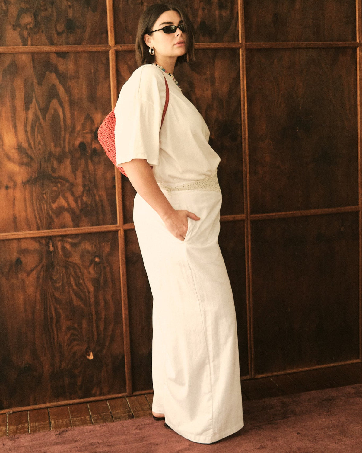 A woman wearing a white short-sleeve maxi dress and black sunglasses stands in profile against a dark wood-paneled wall, holding a red patterned shoulder bag.