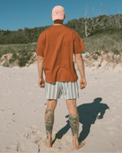 Man walking on a sandy beach wearing a rust-colored shirt and striped shorts.