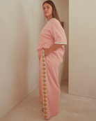 Woman wearing oversized pink shirt and wide-legged pink pants with gold shell detailing down the side. Featuring pockets in the pants. 