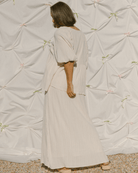 Woman wearing a long, light-colored dress standing against a textured wall.