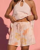 Woman wearing a light pink halter top and floral shorts against a pink background