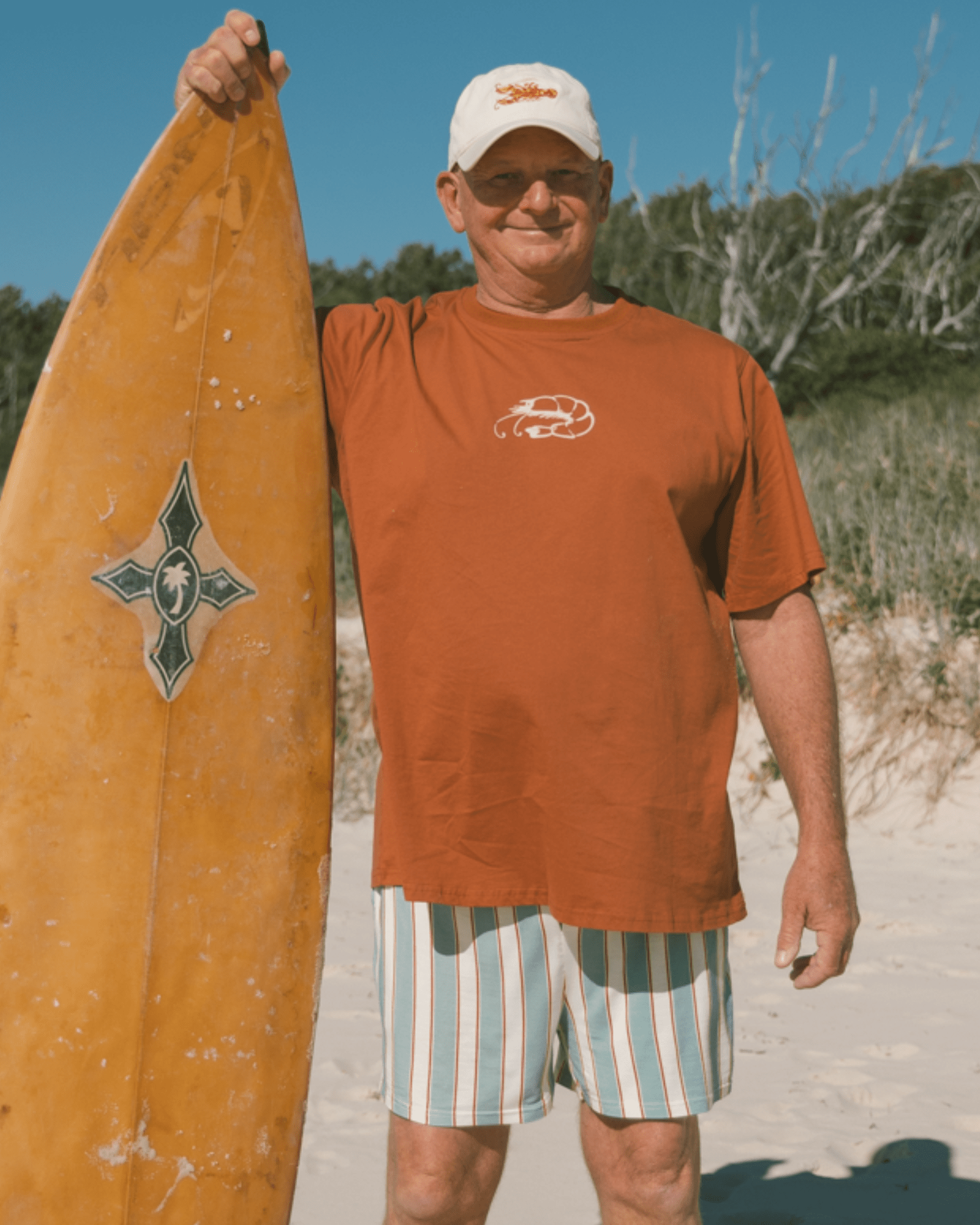 Man holding surfboard and wearing a breathable rust coloured shirt with prawn graphic logo, a beige lobster embroidered hat and blue, beige & rust striped shorts.