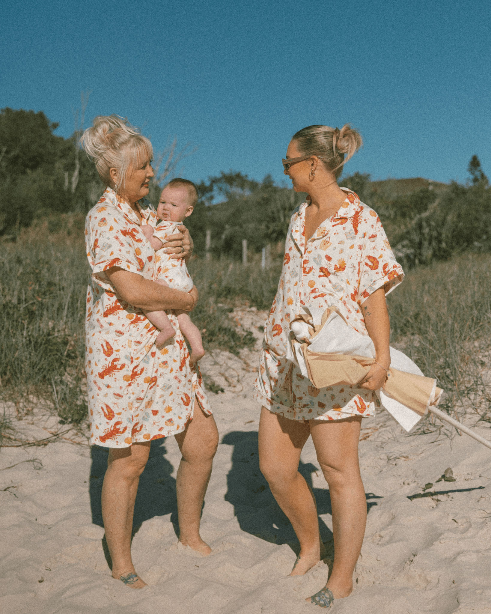Two women in matching Christmas PJ outfits standing on a sandy beach with a baby.