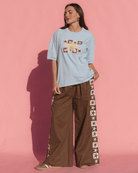 Woman wearing a light blue t-shirt with brown graphic floral & star design and brown pants with star patterns against a pink background