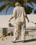 Person wearing a beige top and floral pants walking outdoors with palm trees and a white wall in the background.