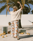 Woman in a casual outfit with floral pants and sunglasses, standing outdoors with palm trees and oranges.