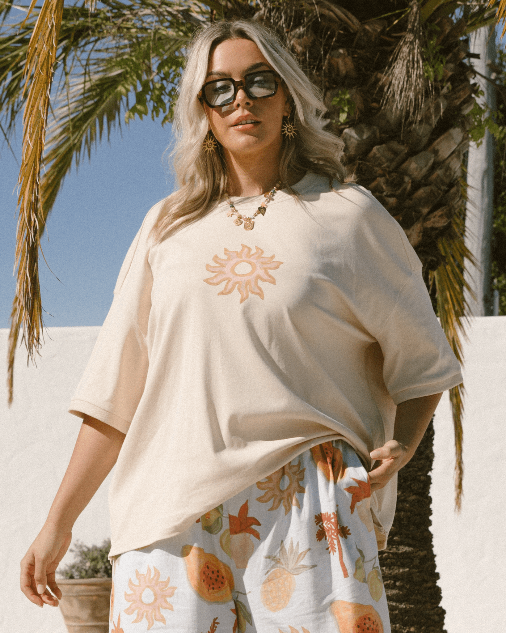 Woman wearing a beige t-shirt with a sun design and floral pants outdoors.