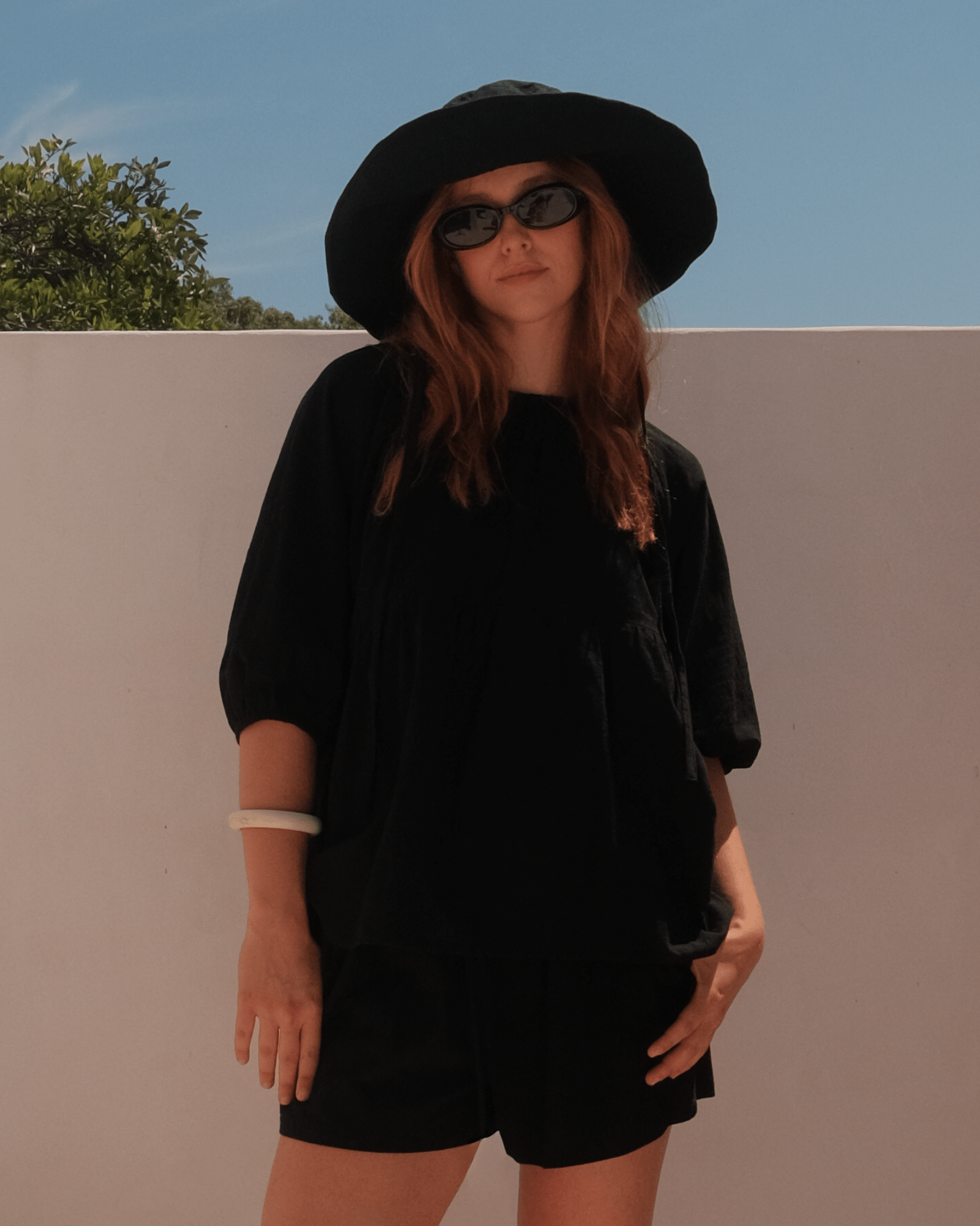 Person wearing a black outfit with a summery hat and sunglasses against a plain background