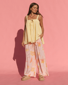 Woman wearing a flowy yellow cotton top and light pink  floral pants against a pink background