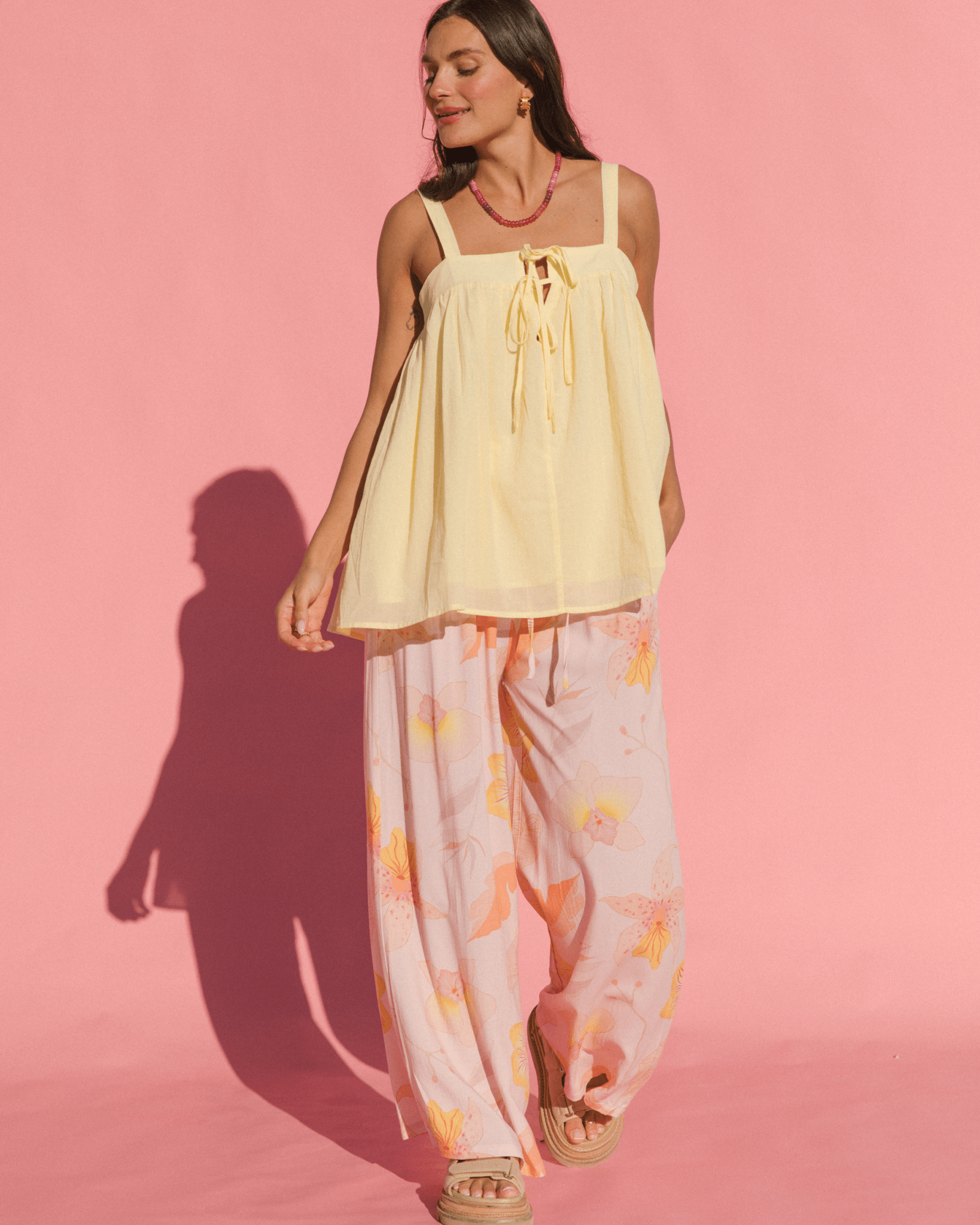 Woman wearing a light yellow top and floral pants against a pink background